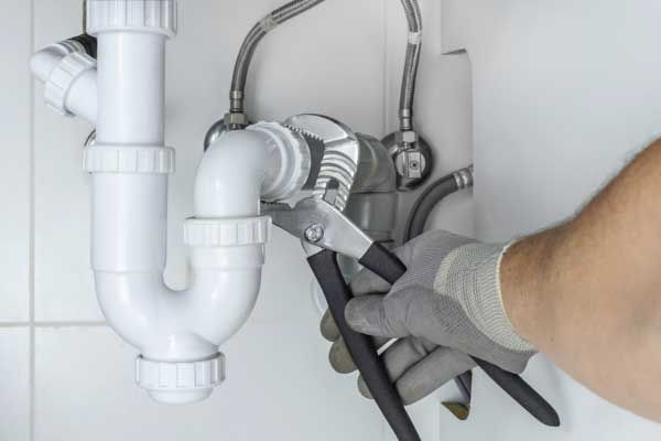 A person wearing gray work gloves uses pliers to tighten a white plastic sink drain pipe under a cabinet.