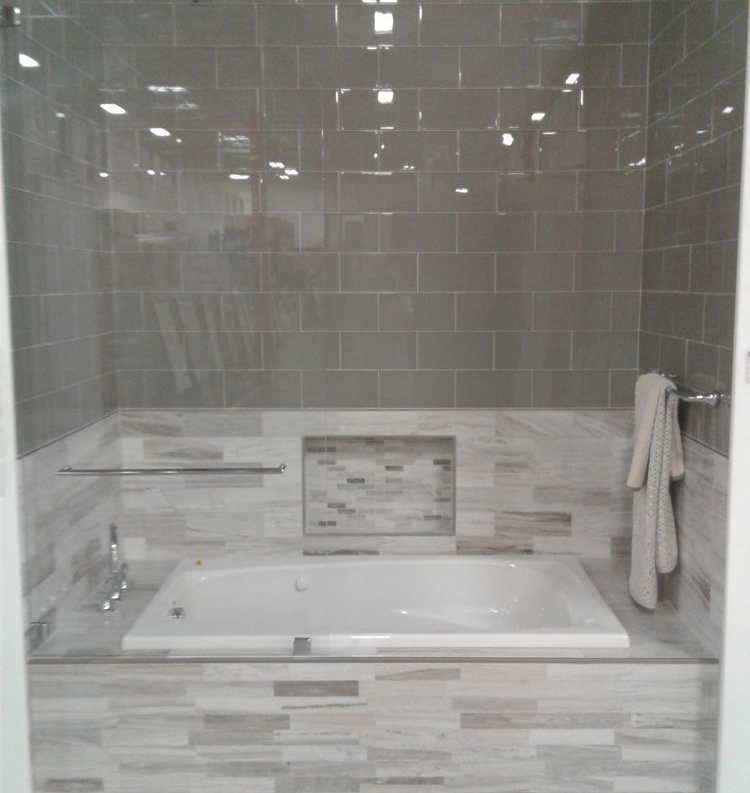 A white soaking tub set against a wall with dark gray subway tiles above a lower section of light, horizontal stone tiles.