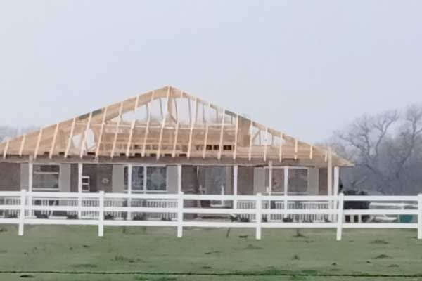 A house under construction with exposed wooden roof trusses, set behind a white vinyl fence in a grassy field.