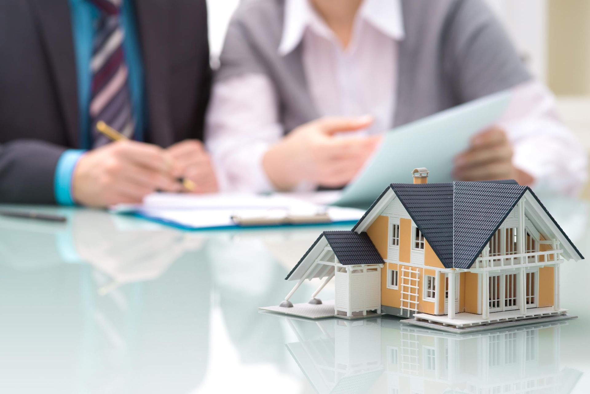 Miniature house on a table with people reviewing documents in the blurred background.