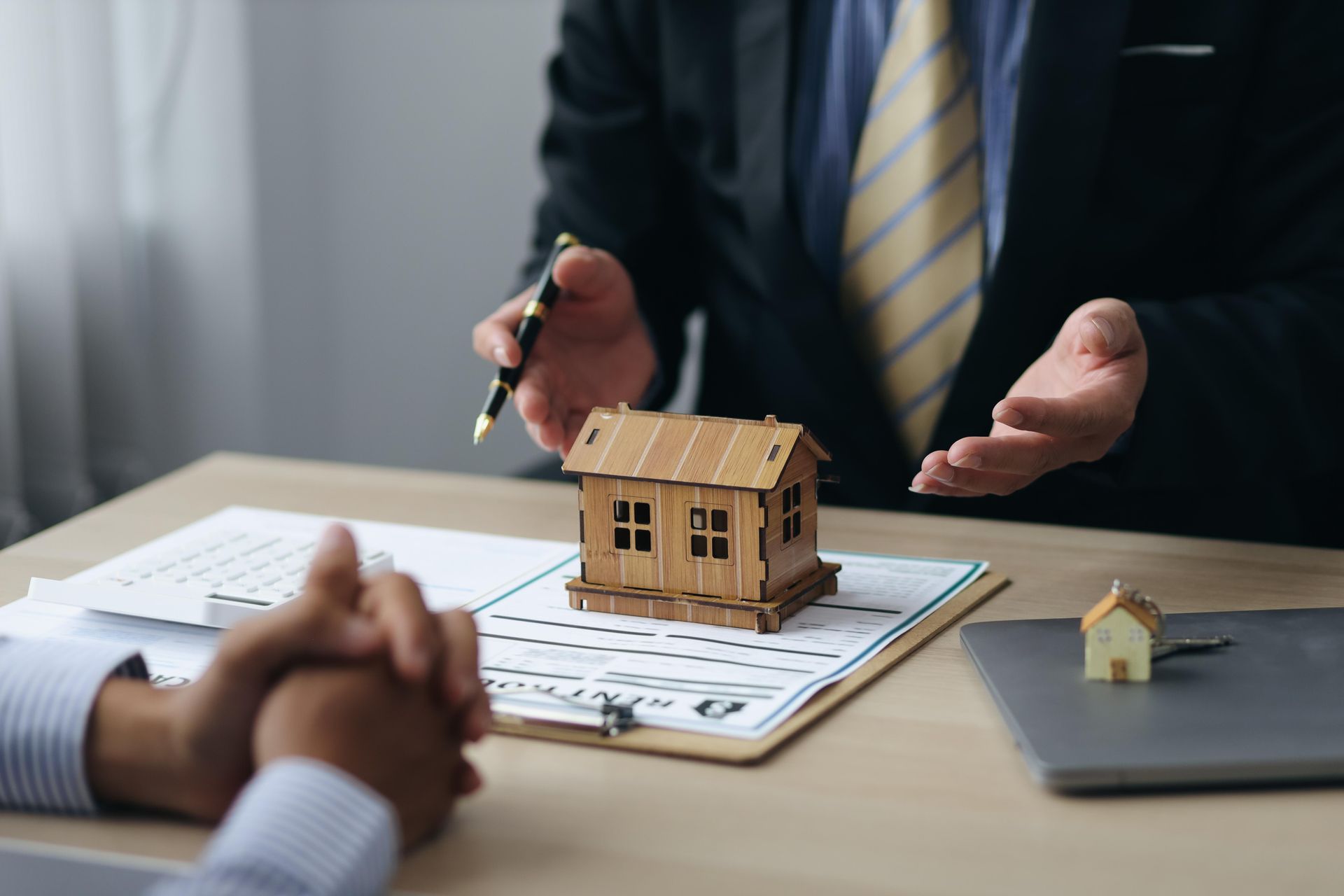 Real estate agent gesturing toward a small wooden house model on a desk. Documents, laptop, and another model house present.