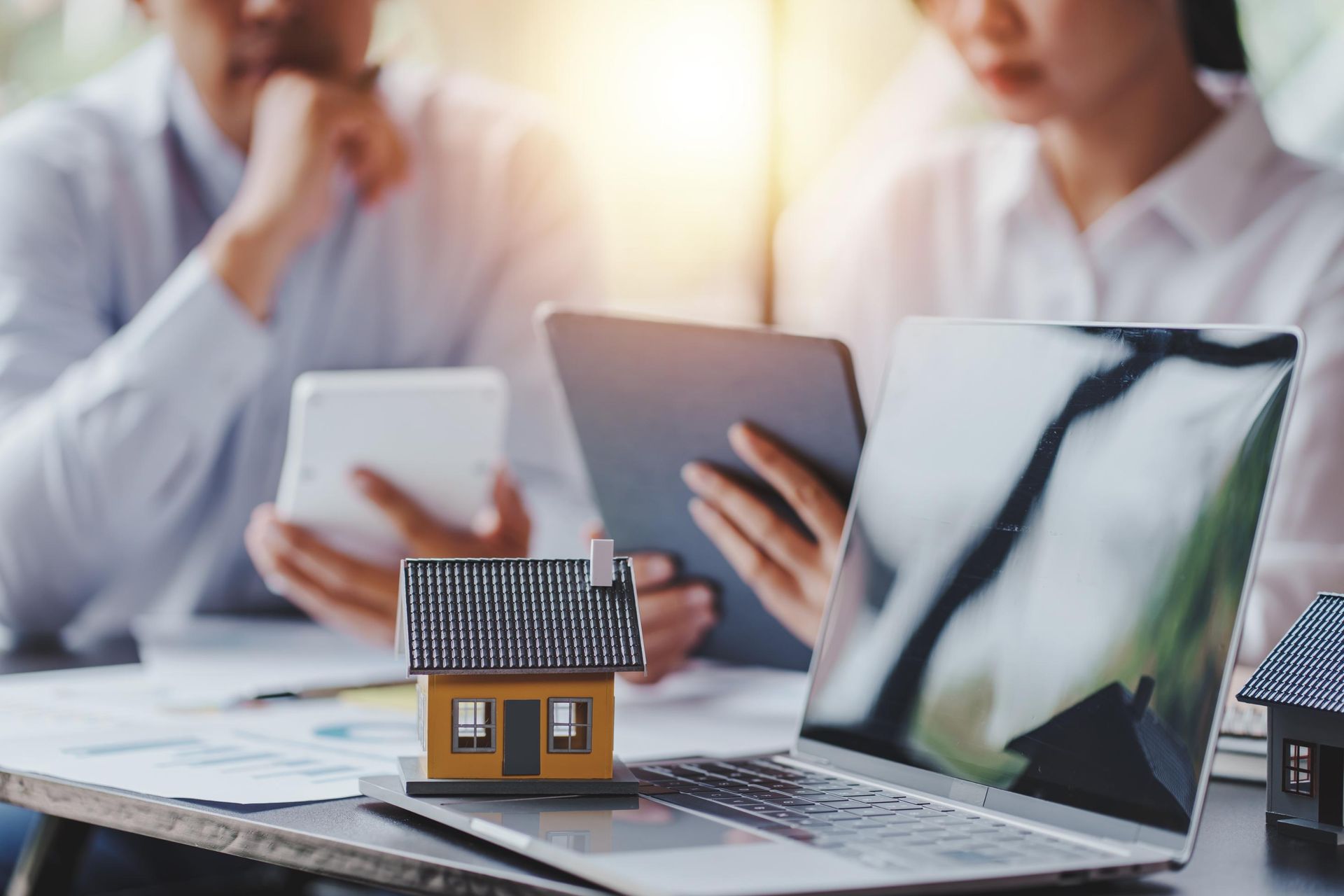 Two people reviewing documents, laptop, and small house model on a table.