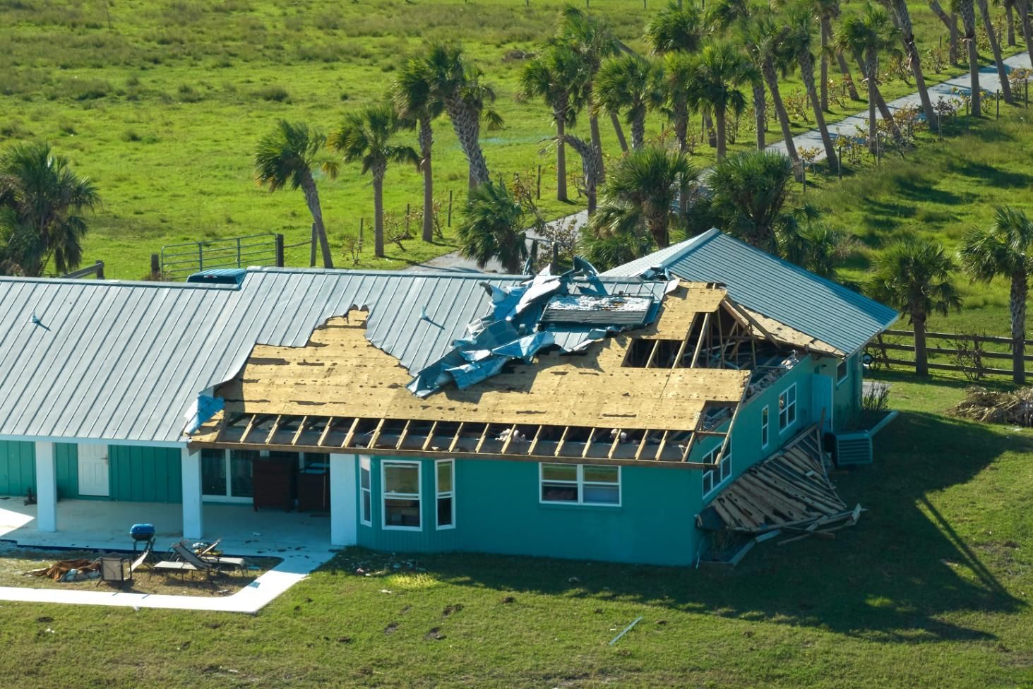 House with roof damage from a storm, blue siding, green lawn, palm trees.