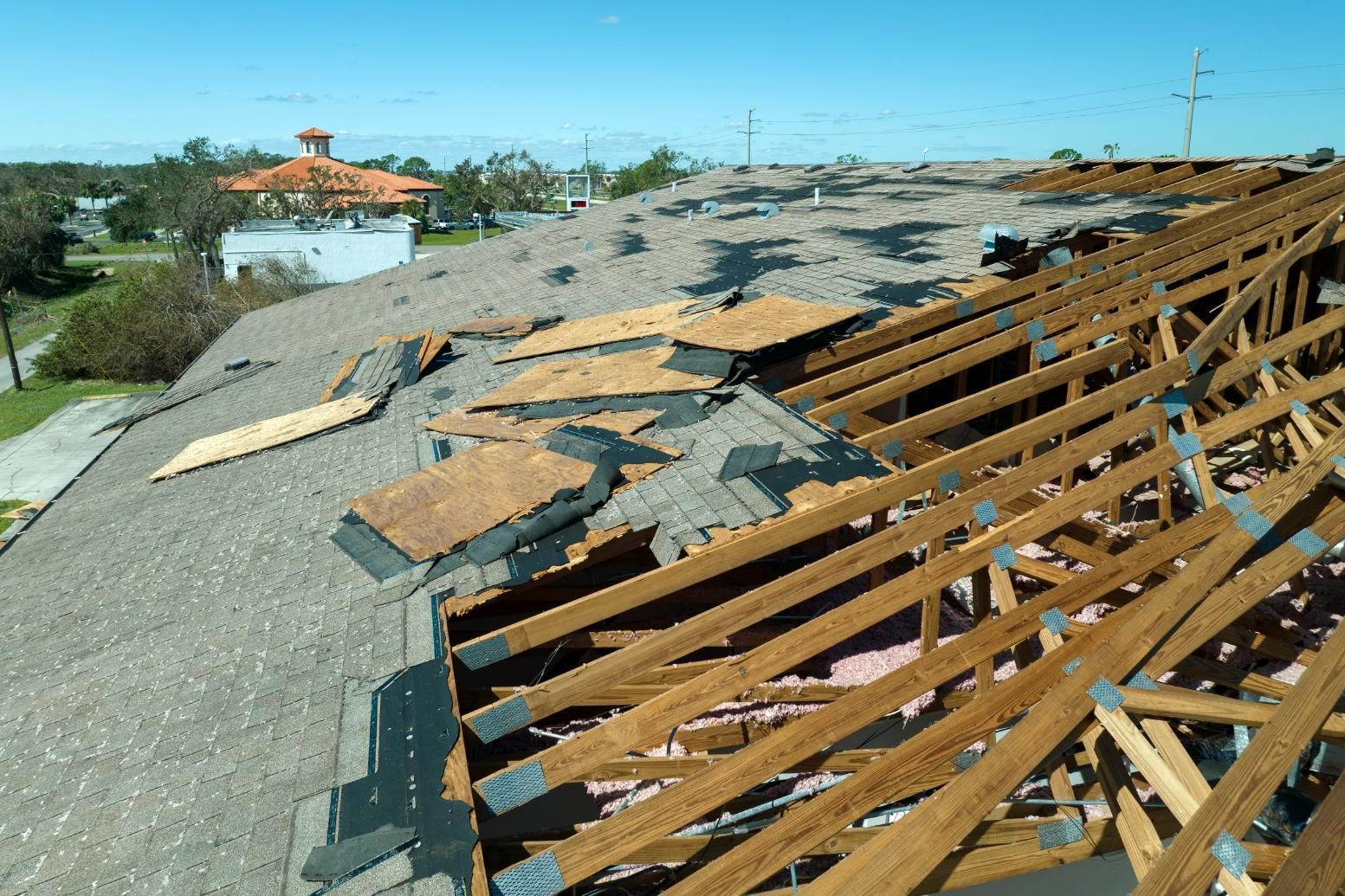 Damaged roof with exposed wooden beams, brown insulation, and blue sky.