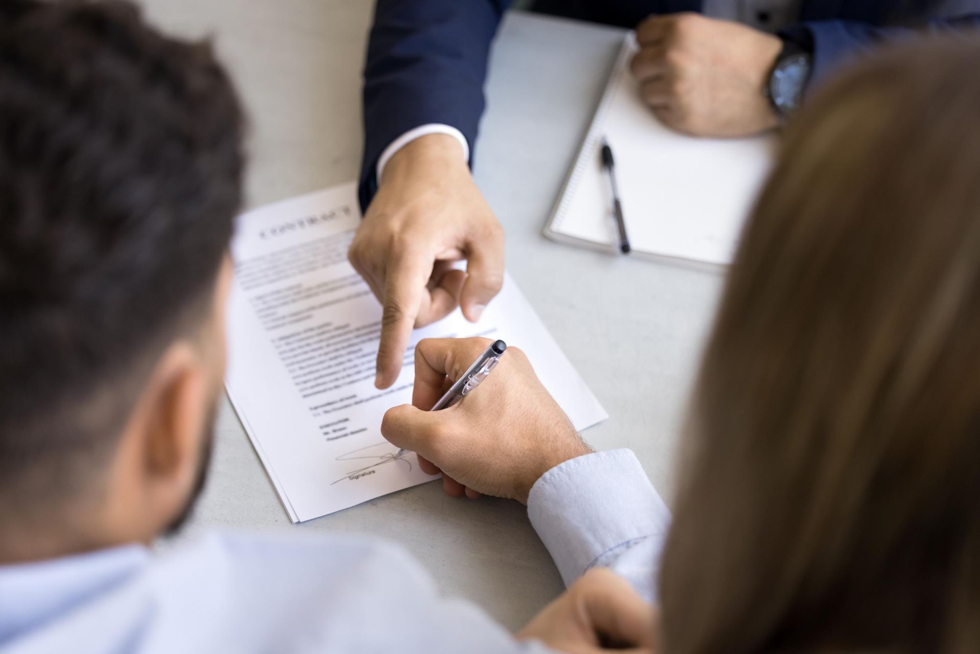 Person signing a document, being pointed to by another person; two others watch.