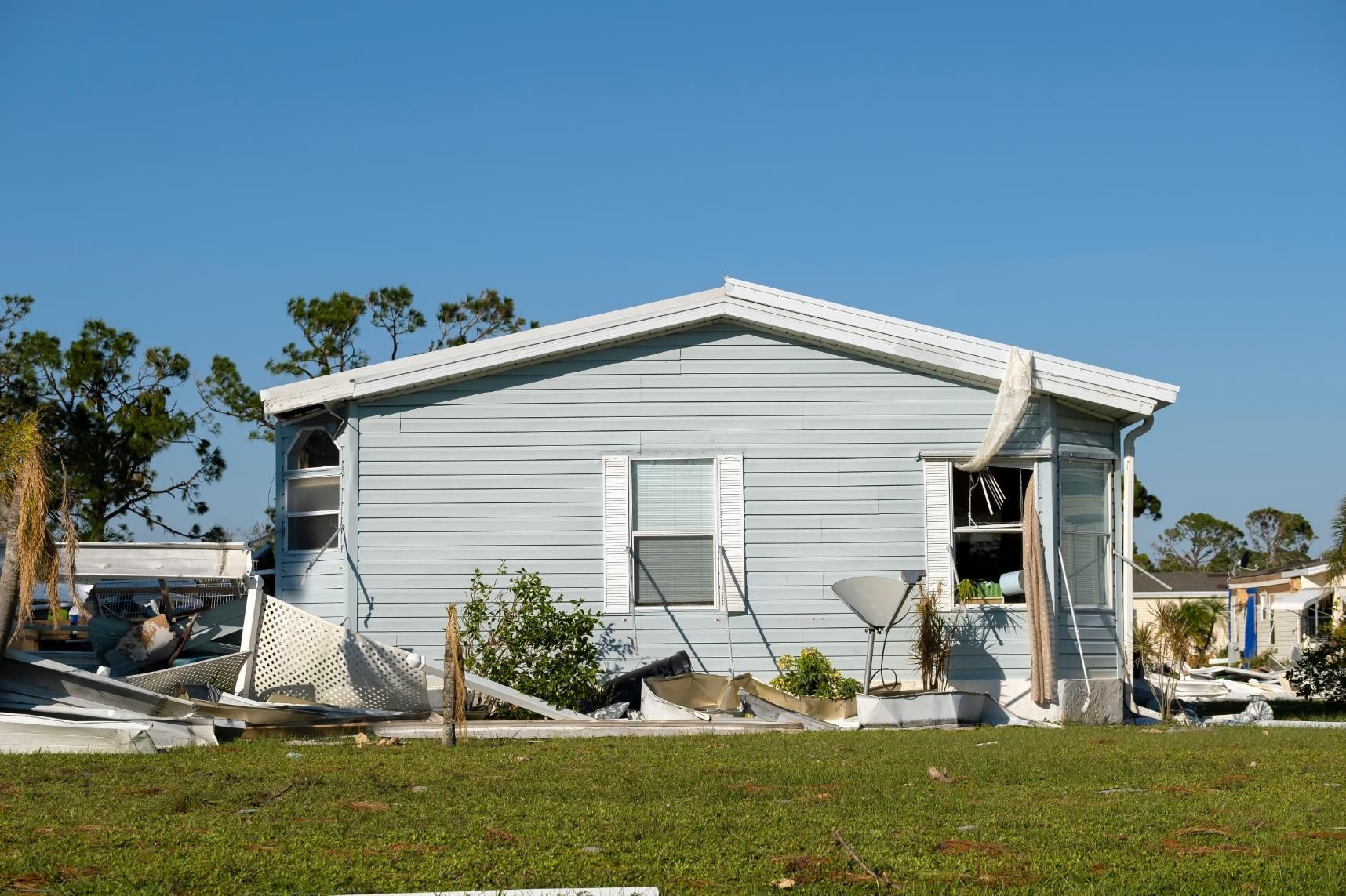 Damaged blue mobile home with broken windows, debris, and satellite dish on a grassy field under a clear sky.