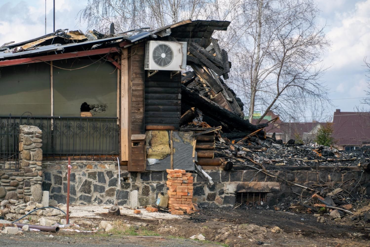Charred remains of a house after a fire, with visible damage to the roof, walls, and surrounding area.