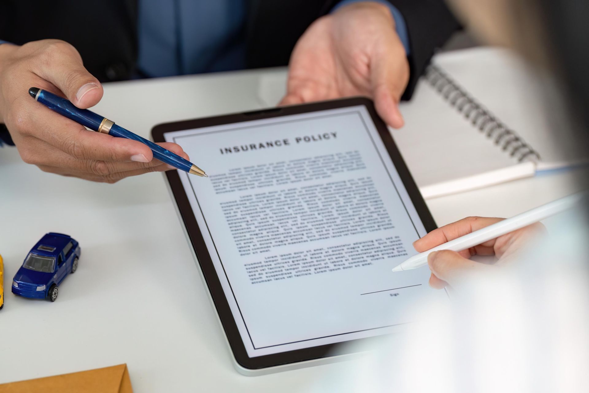 Hands pointing at and signing an insurance policy on a tablet. Includes a toy car and notepad.
