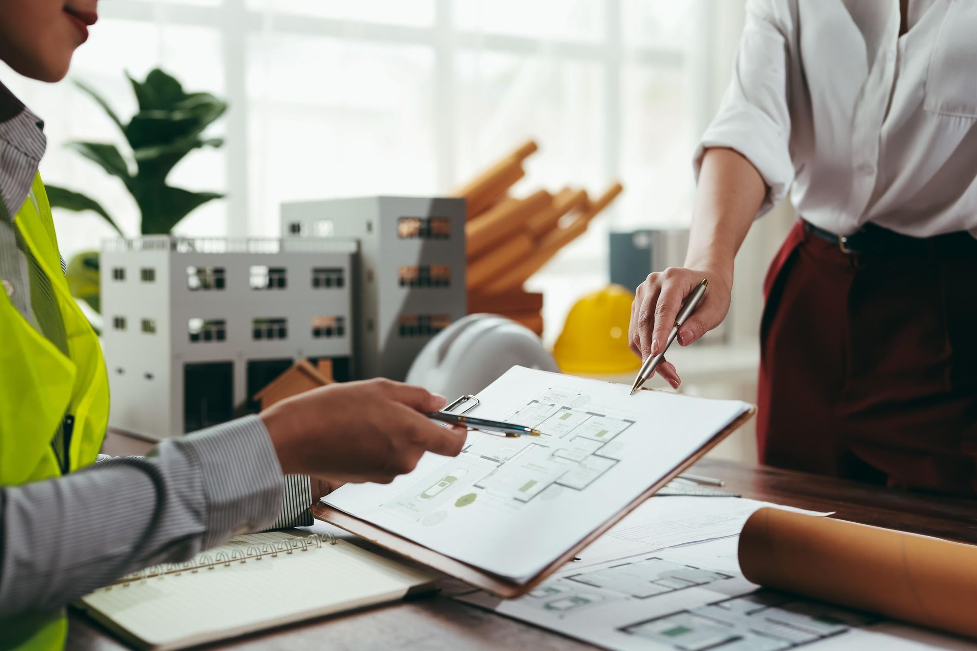 Two people reviewing architectural plans, with building models and construction materials on a desk.