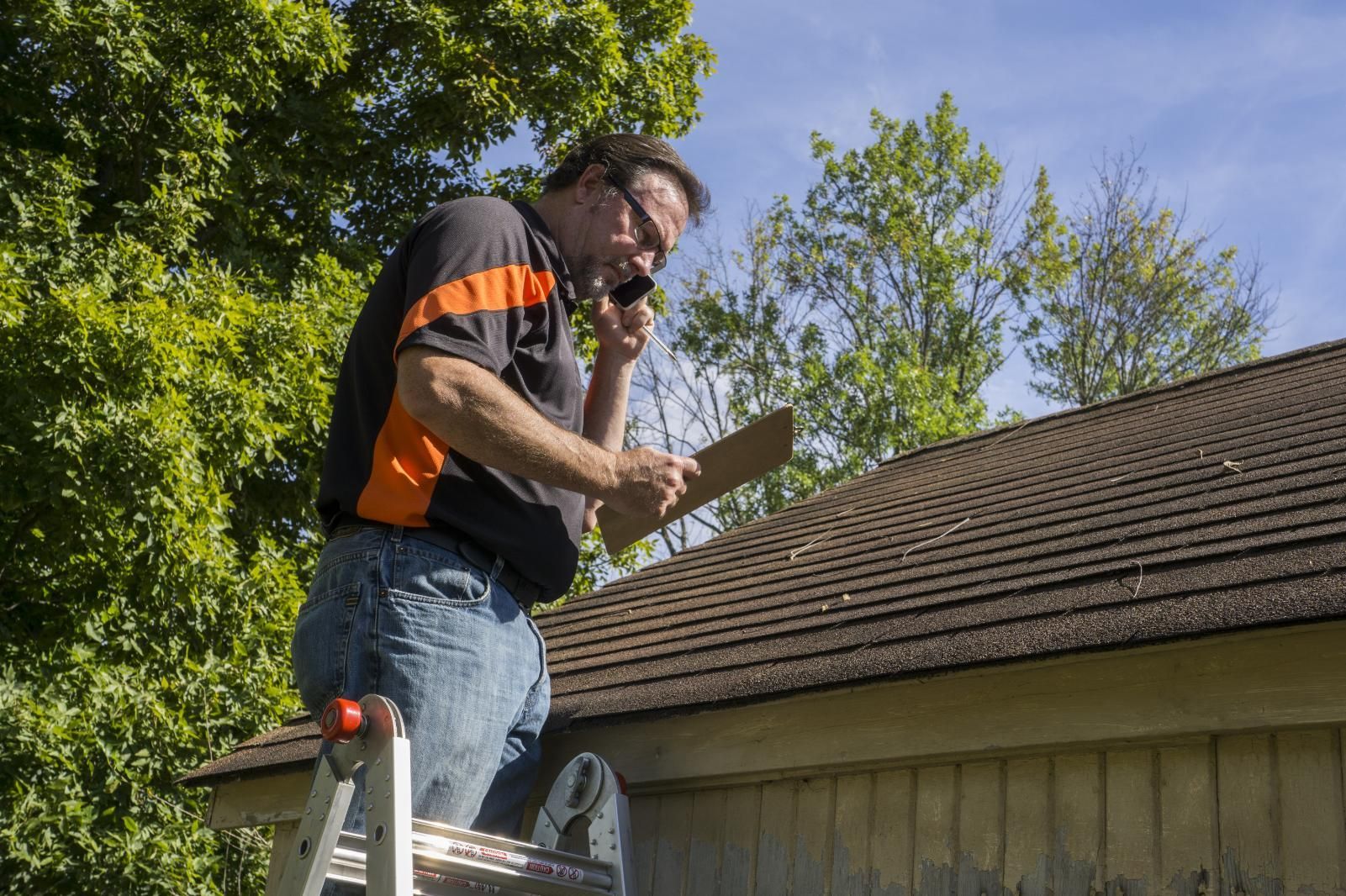 Man on a ladder inspecting a roof, talking on the phone, holding a clipboard, sunny outdoor setting.
