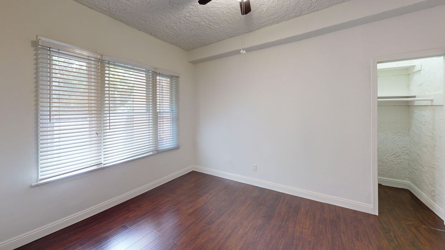 Empty room with dark wood floor, white walls, window with blinds, and closet.