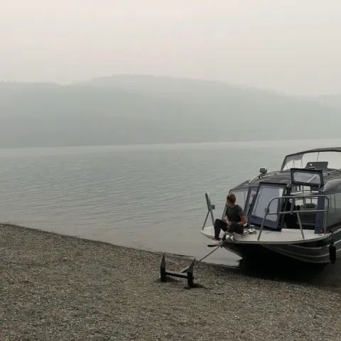 A man is sitting on a boat on the shore of a lake.