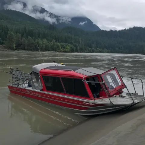 A red boat is docked on the shore of a river with mountains in the background.