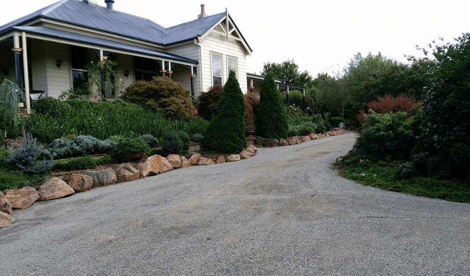 A driveway leading to a large white house surrounded by trees and rocks.