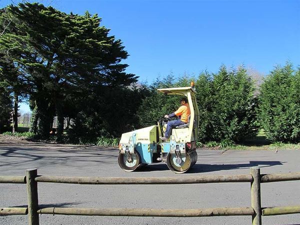 A man is driving a roller on a road next to a wooden fence