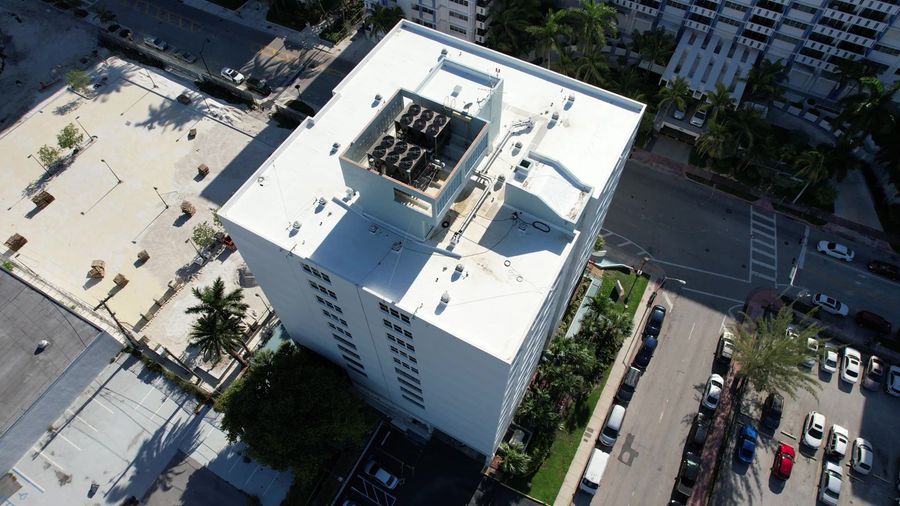 Aerial view of a white high-rise building with a flat roof, surrounded by parked cars and trees in an urban setting.