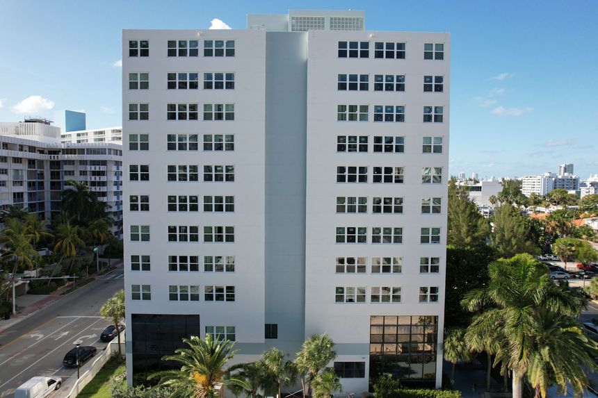 White high-rise building with rectangular windows, blue-gray center facade, and palm trees in foreground.