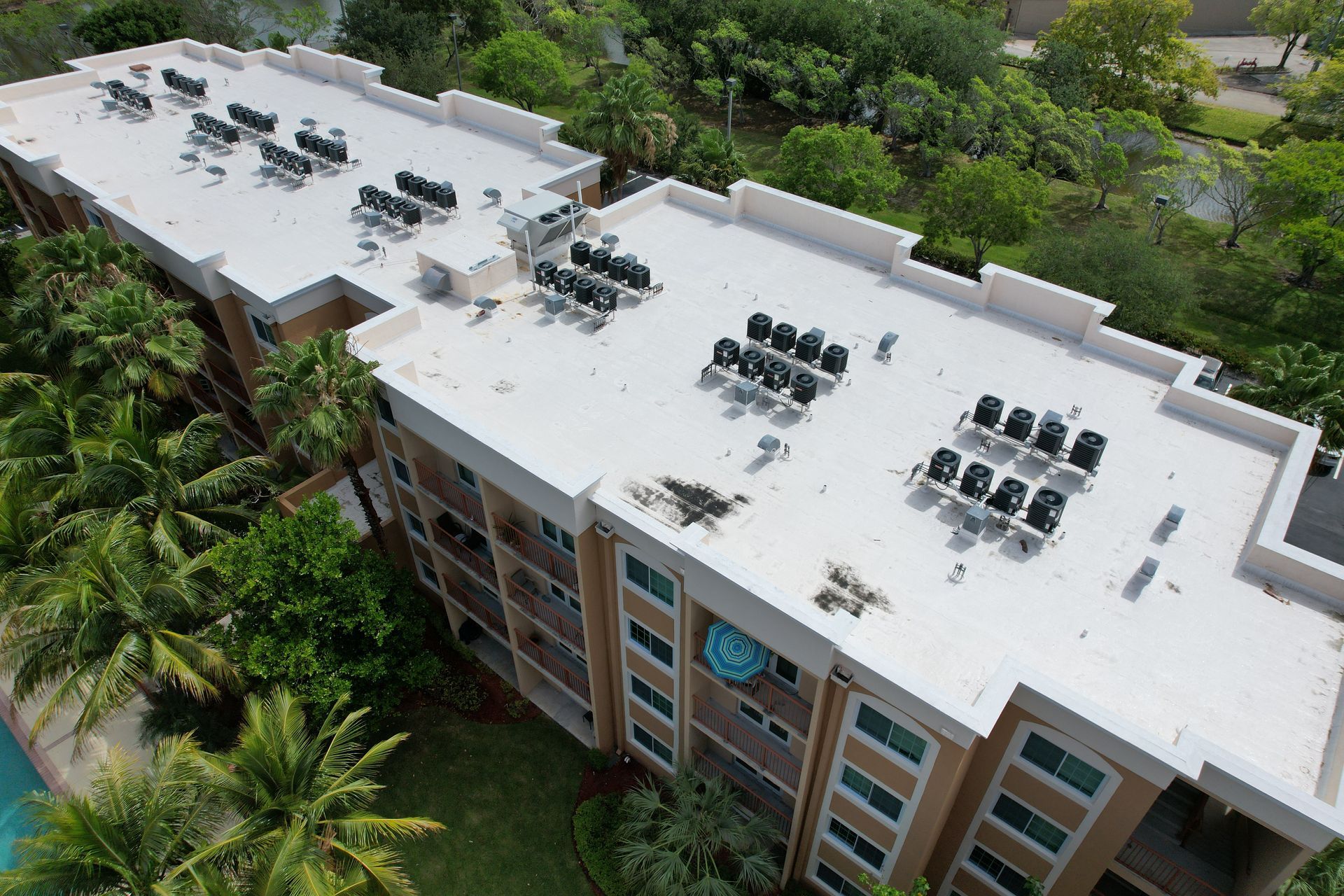 Aerial view of a multi-story building with a flat white roof covered in HVAC units, surrounded by lush green trees.