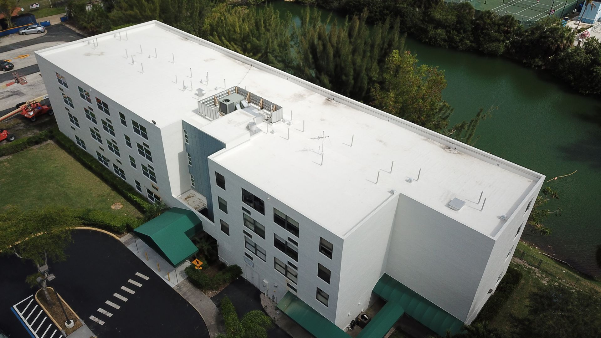 Aerial view of a white building with multiple stories and a flat roof, near a green body of water.