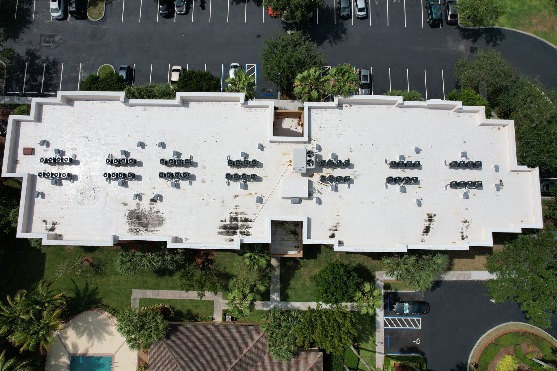 Aerial view of a white flat roof with multiple HVAC units; surrounded by trees, parking lot and a small pool.
