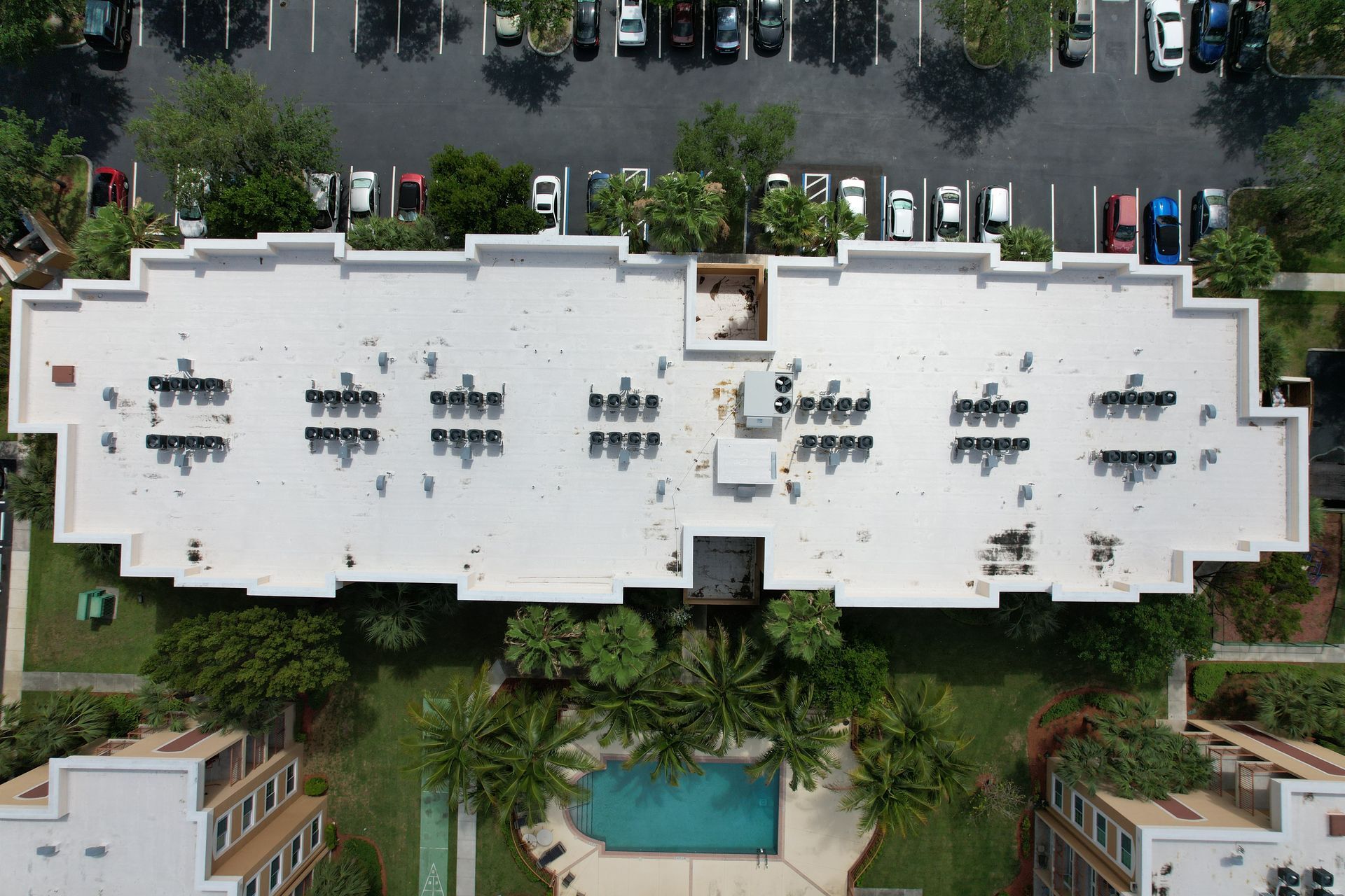 Overhead view of a white building with rooftop air conditioning units, parking lot, and a swimming pool.