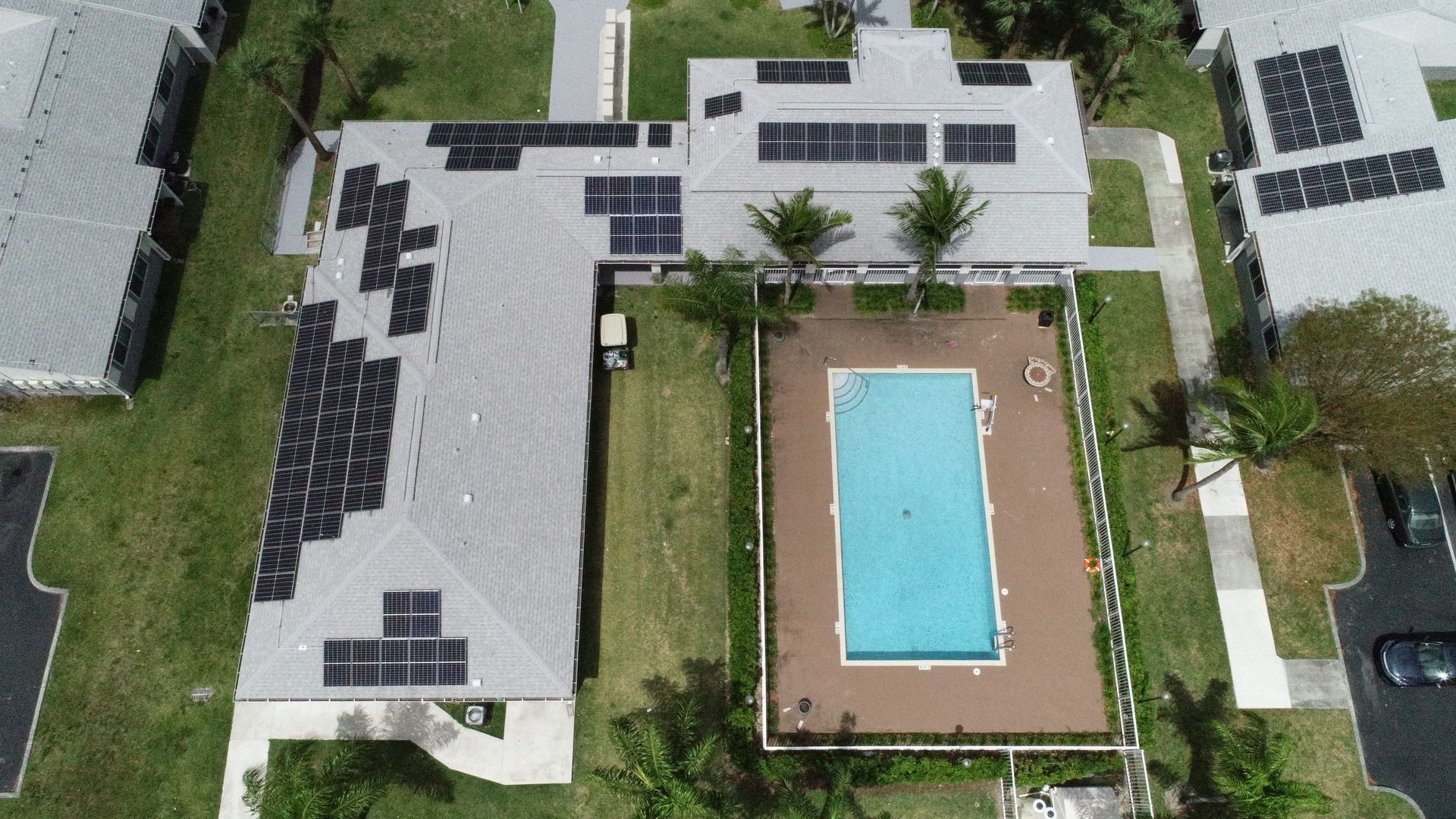 Aerial view of a building with solar panels, pool in the center, and green lawn around it.