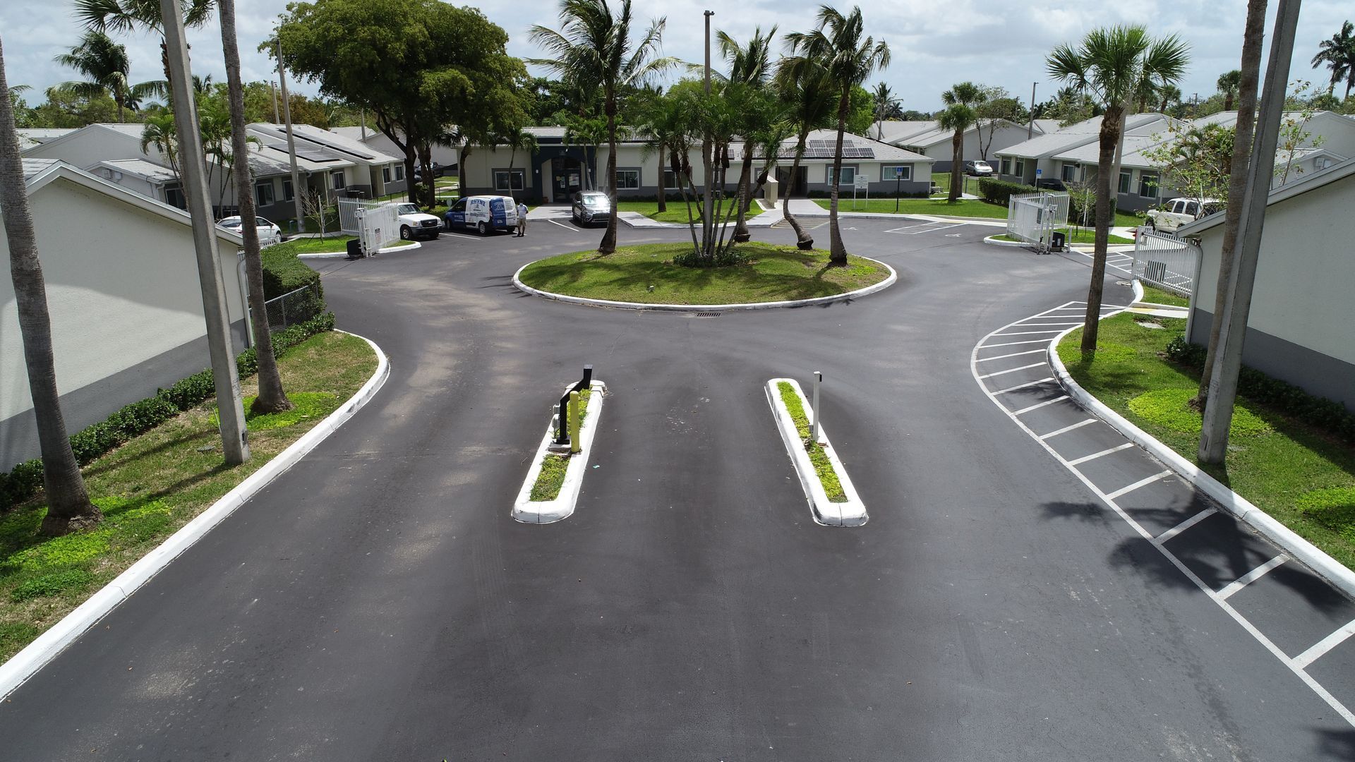 Road entrance to a community with a roundabout, palm trees, and white houses.