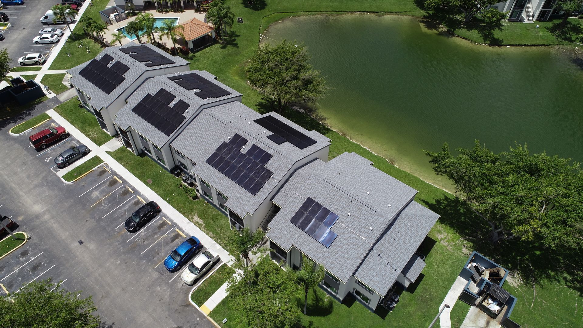 Aerial view of a residential building with solar panels on its roof next to a parking lot and a lake.