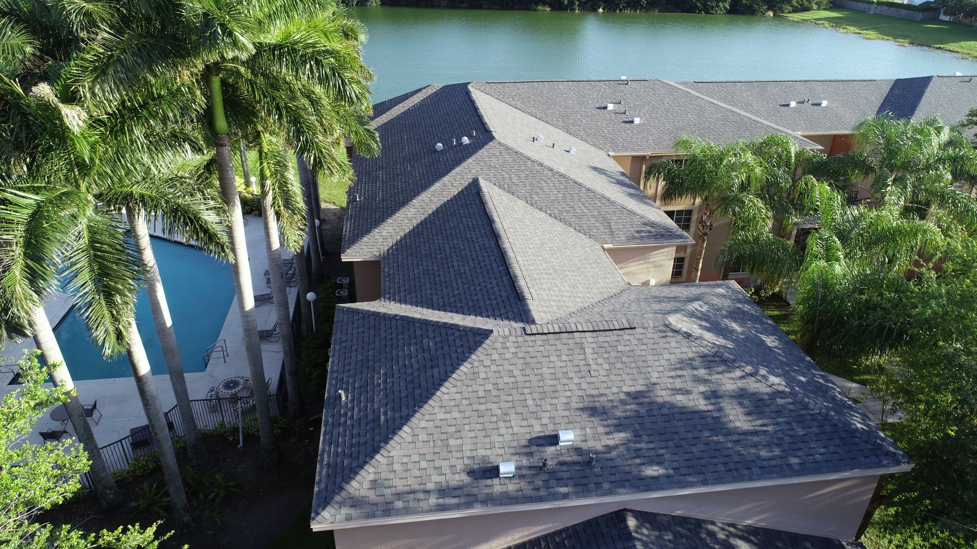 Aerial view of homes with dark gray roofs near a pool and lake, surrounded by palm trees.