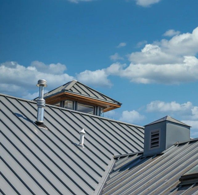 Gray metal roof with skylight and chimneys against a blue sky with puffy white clouds.