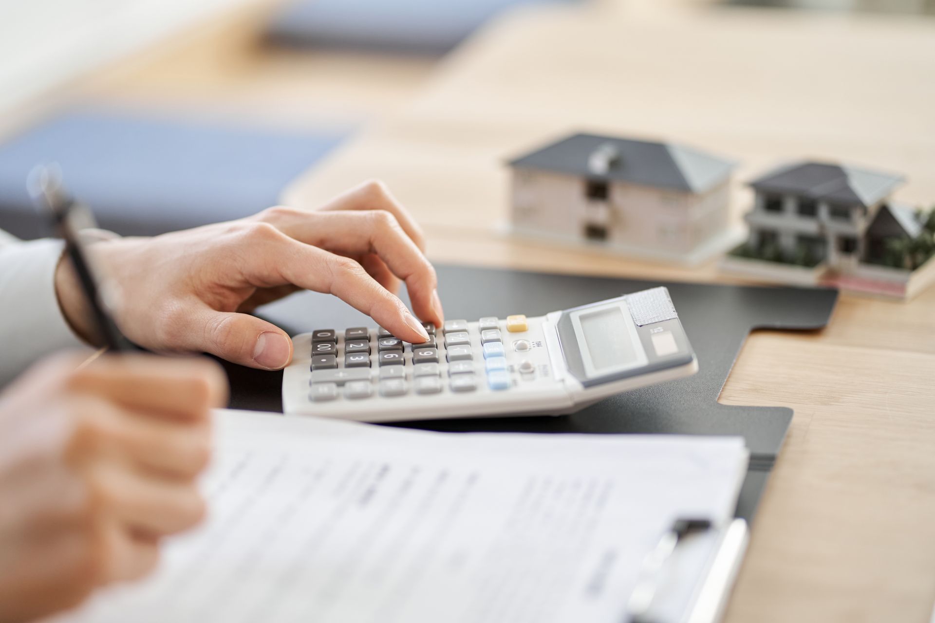 Hands using a calculator, calculating real estate costs, with house models and paperwork.