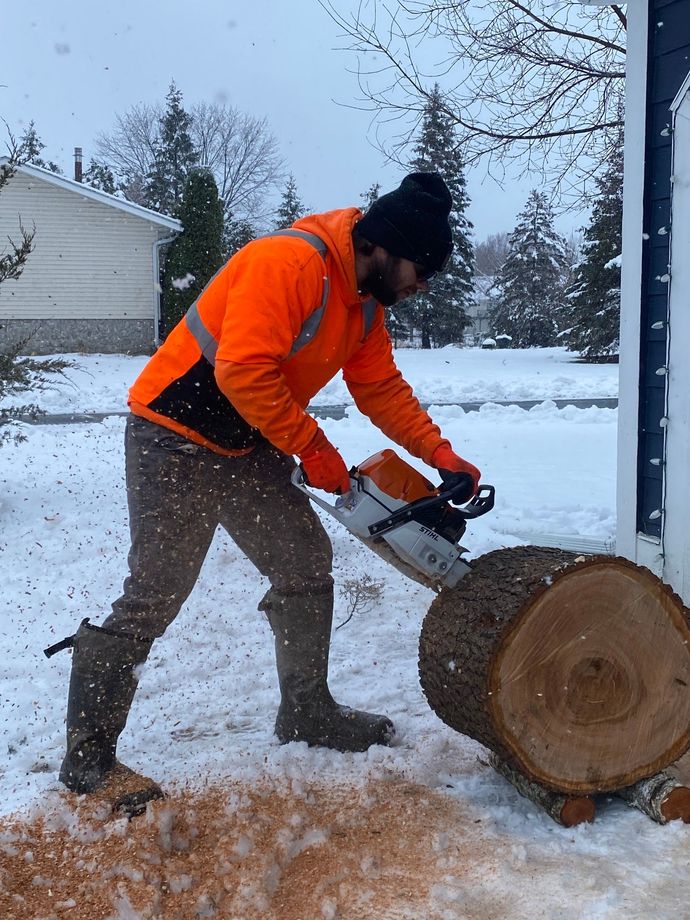Man in orange jacket using a chainsaw on a log in a snowy setting.