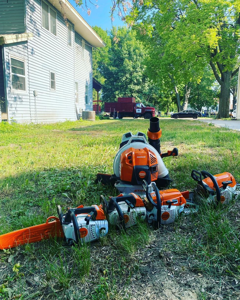 A collection of orange and white chainsaws and a leaf blower lie on a grassy lawn near a house.