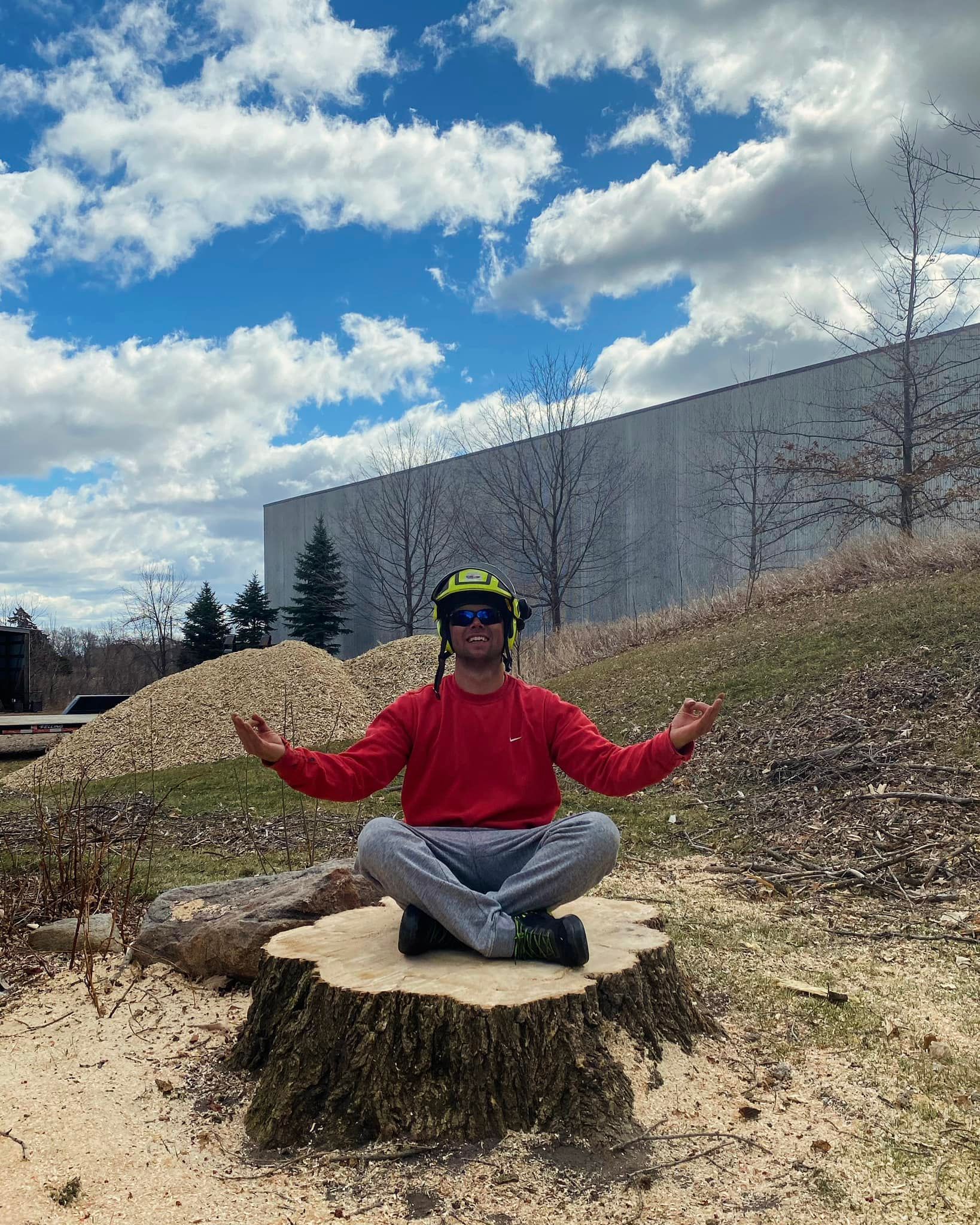 Man in red sweater meditates on a tree stump outdoors, wearing safety gear; blue sky background.