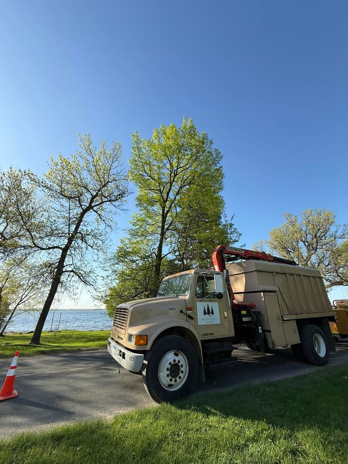 Tan dump truck parked near a lake with trees and blue sky.