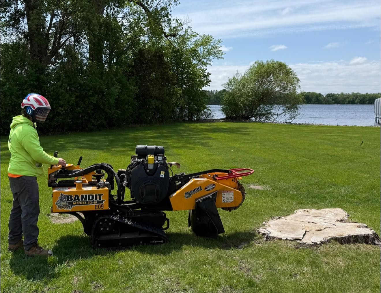 Person grinding a tree stump with a Bandit stump grinder on a grassy lawn by a lake.