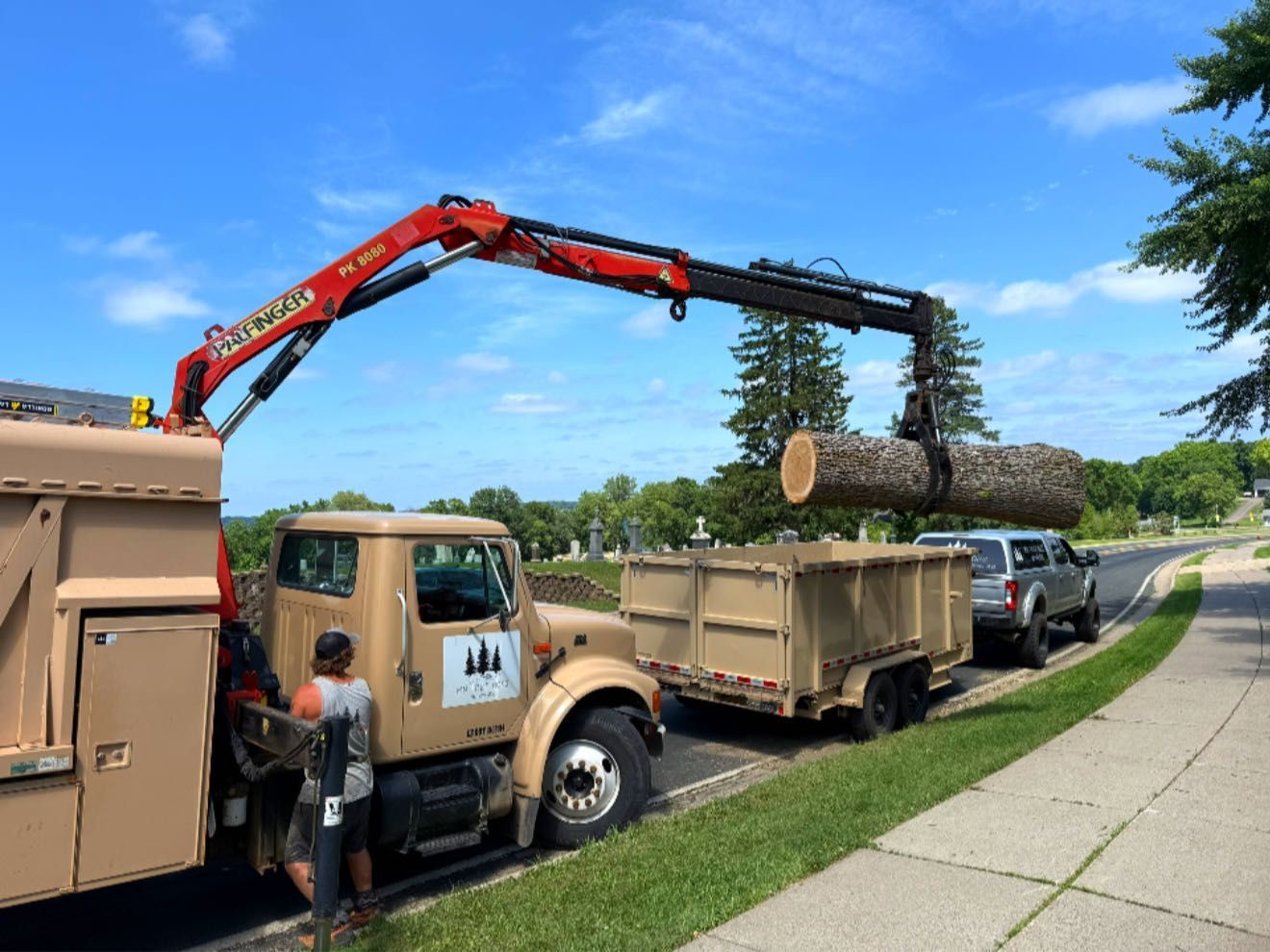 A truck with a crane lifting a large log into a trailer on a sunny roadside.