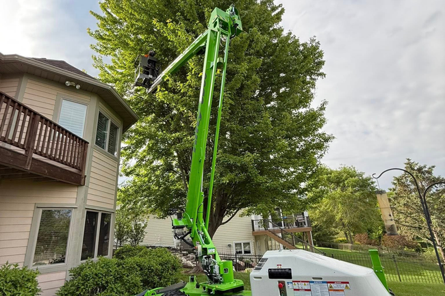 A green tree-trimming lift extends towards a large tree, next to a house with a balcony.