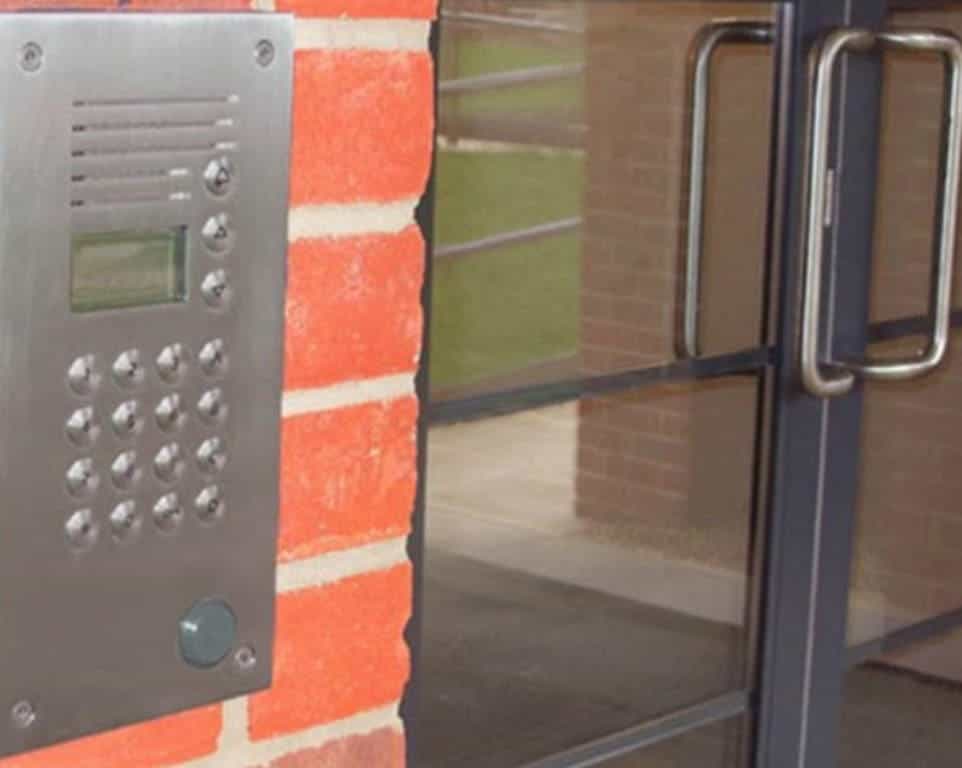 Metal intercom on red brick wall next to glass door with silver handles.