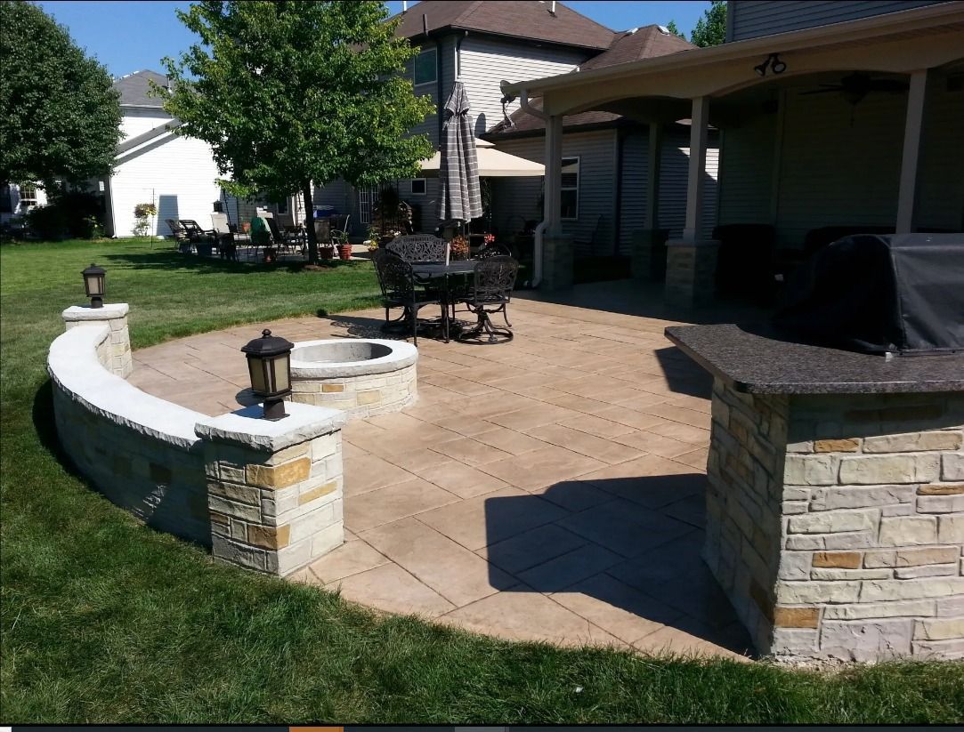 Backyard patio with fire pit, seating, and built-in grill, surrounded by lawn and stone walls.