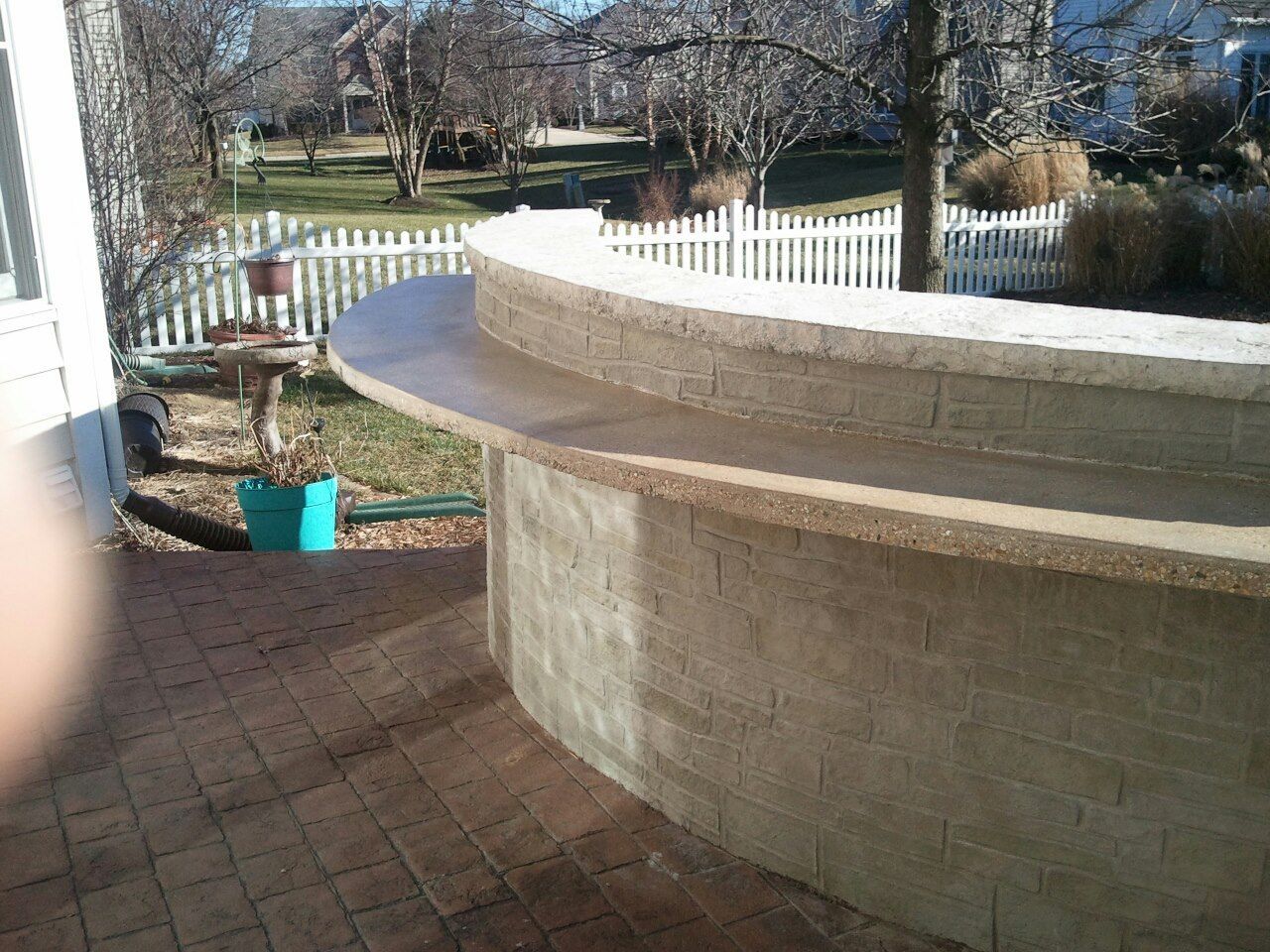 Curved outdoor bar made of light-colored brick. Brown countertop, brick patio, white picket fence.