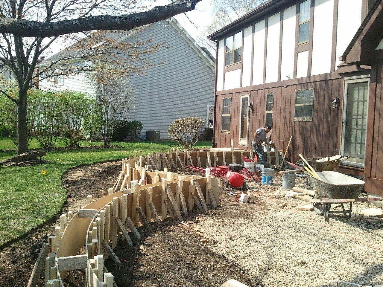 Construction site: wooden forms for a curved concrete structure in a yard. Man working, house in background.