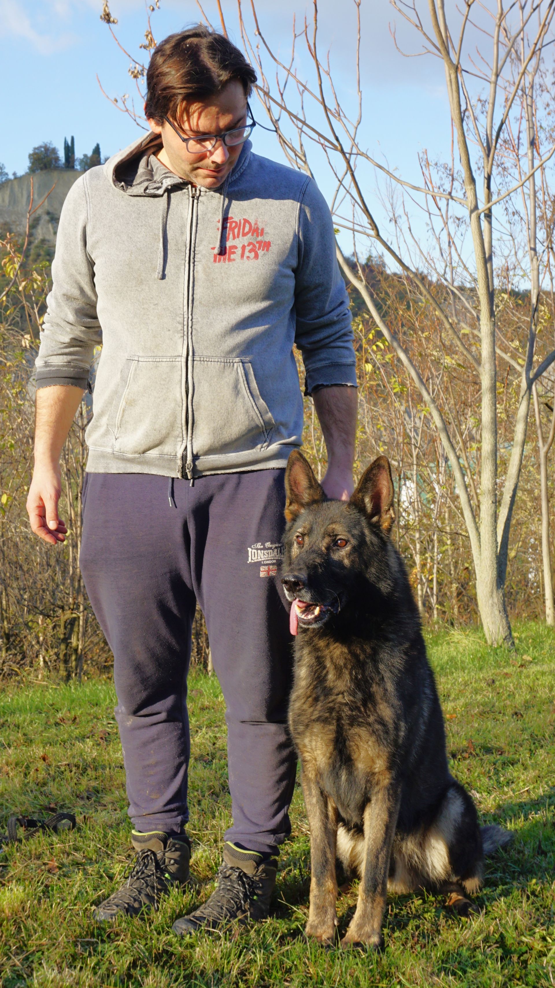 Un uomo con una felpa grigia con cappuccio è in piedi all'aperto con un pastore tedesco tigrato.