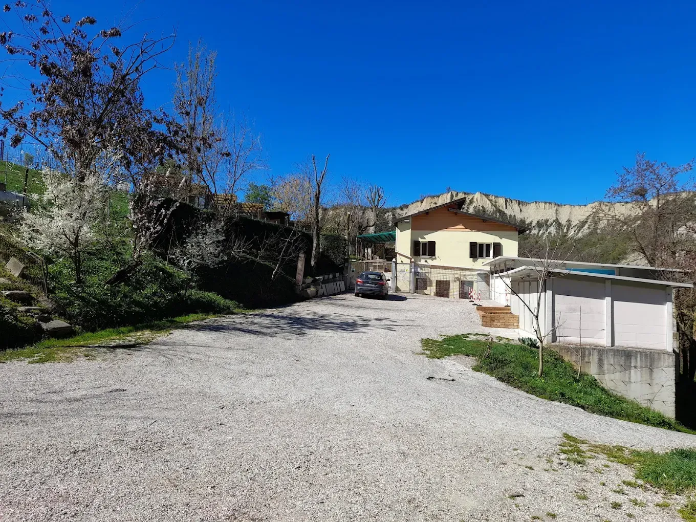Parcheggio in ghiaia di fronte a una casa gialla, sotto un cielo azzurro e limpido. Montagne sullo sfondo.