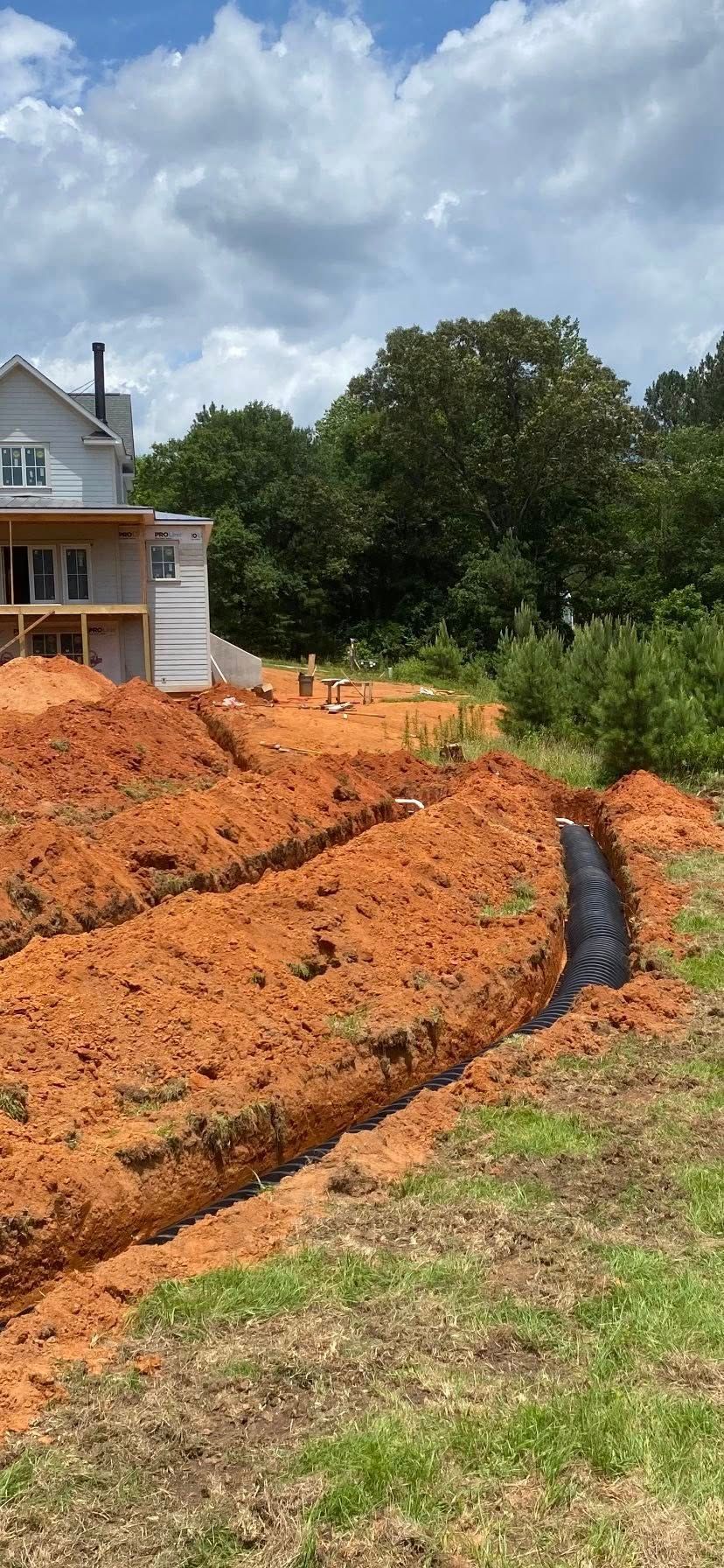 Trenches dug in red earth for a construction project, with a partially-built house in the background under a cloudy sky.