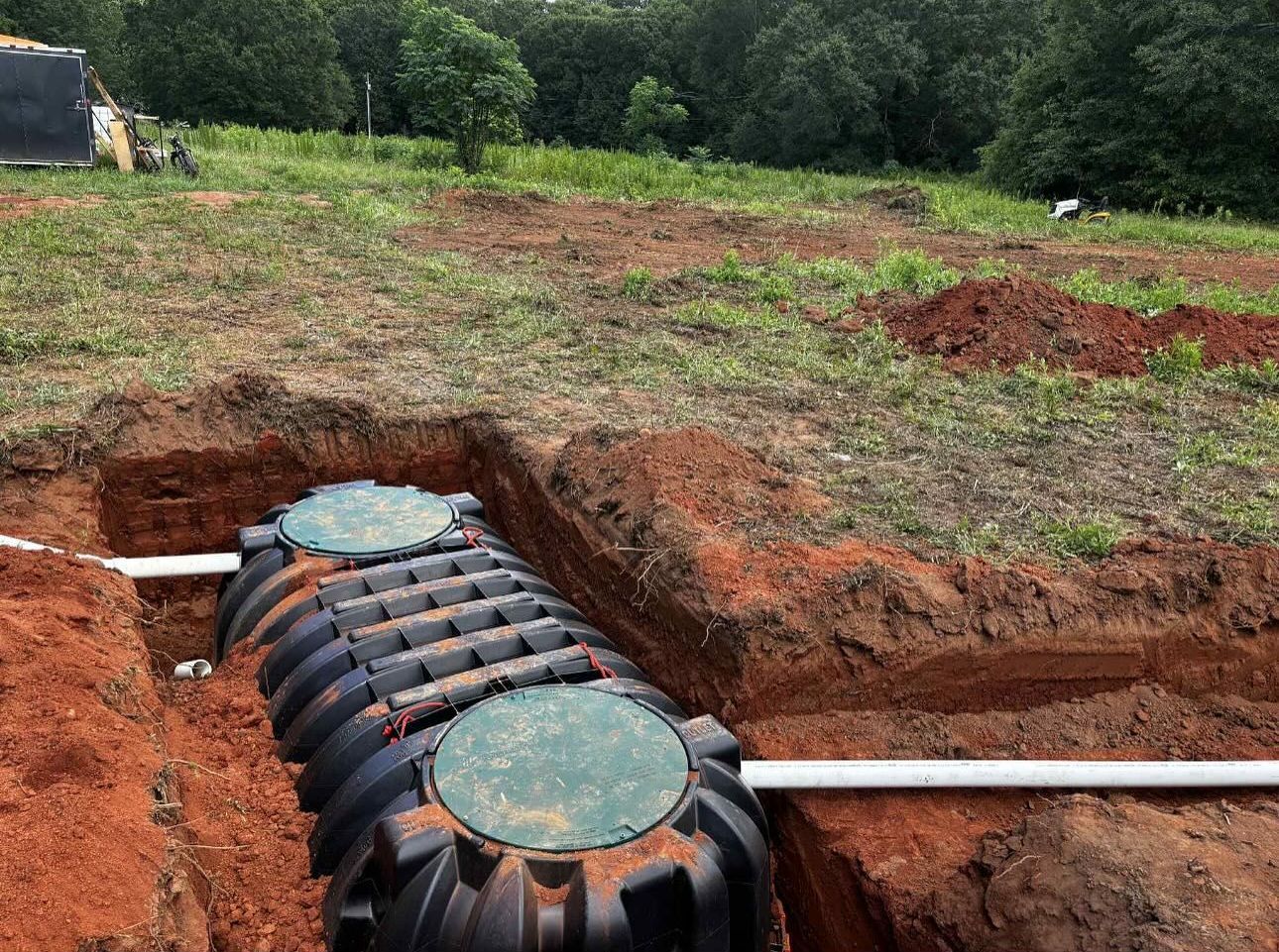 Black septic tank with two green lids sits in a dirt trench, with white pipes connected. Reddish-brown soil surrounds the tank, and green grass is in the background.