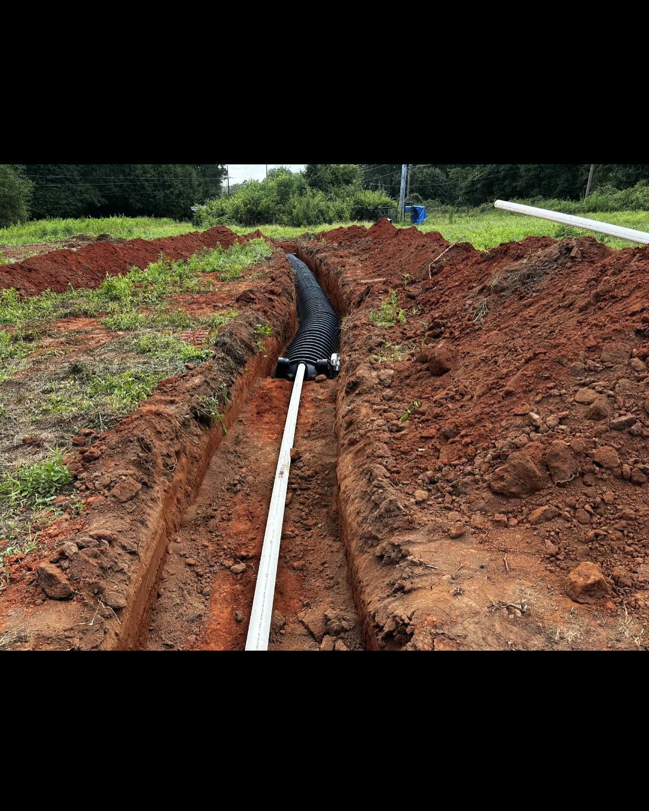 A trench dug in red soil contains a white pipe and a black drainage system, outdoors.