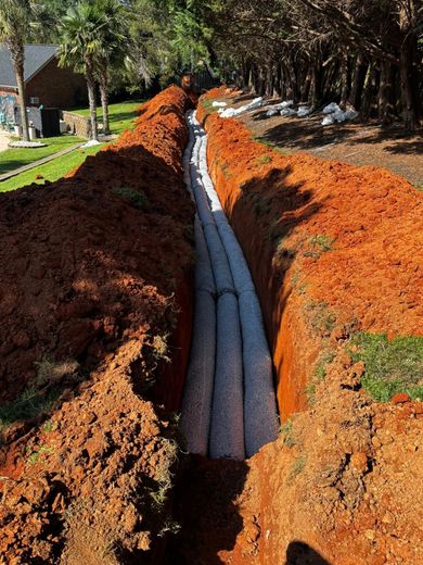 Long trench with several gray drainage tubes installed; surrounded by reddish-brown soil, in a residential yard.