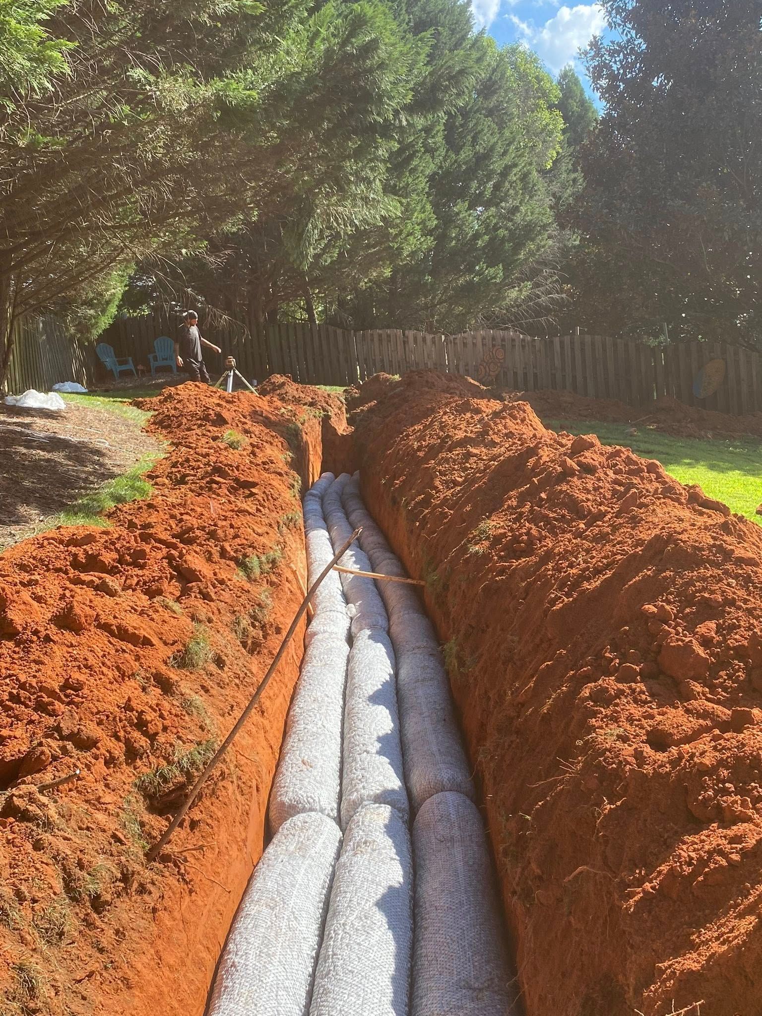 A trench in a yard, lined with gray filter fabric and gravel, ready for drainage pipes. Red clay surrounds the trench.