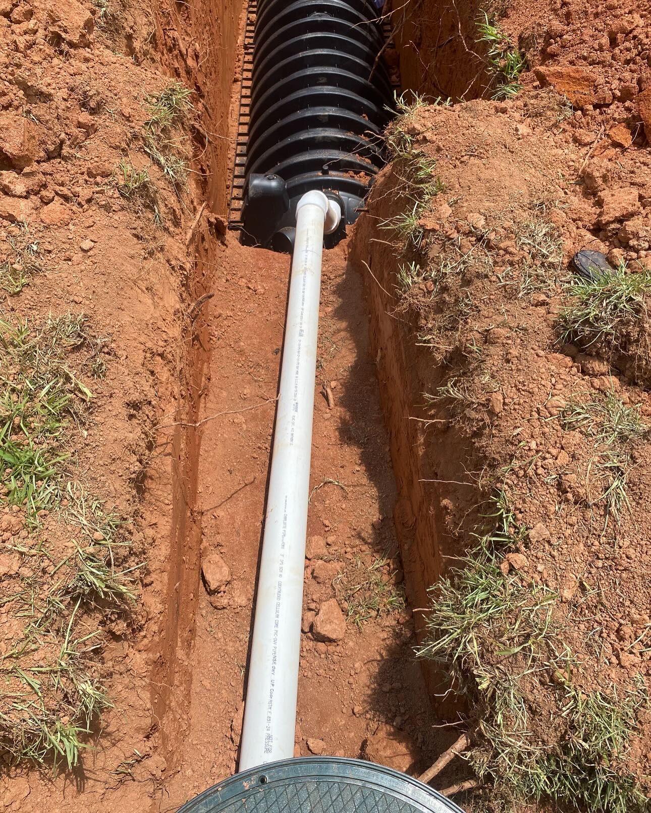 Drainage pipe and corrugated tank installed in a narrow trench in red soil, with grass visible on the edges.